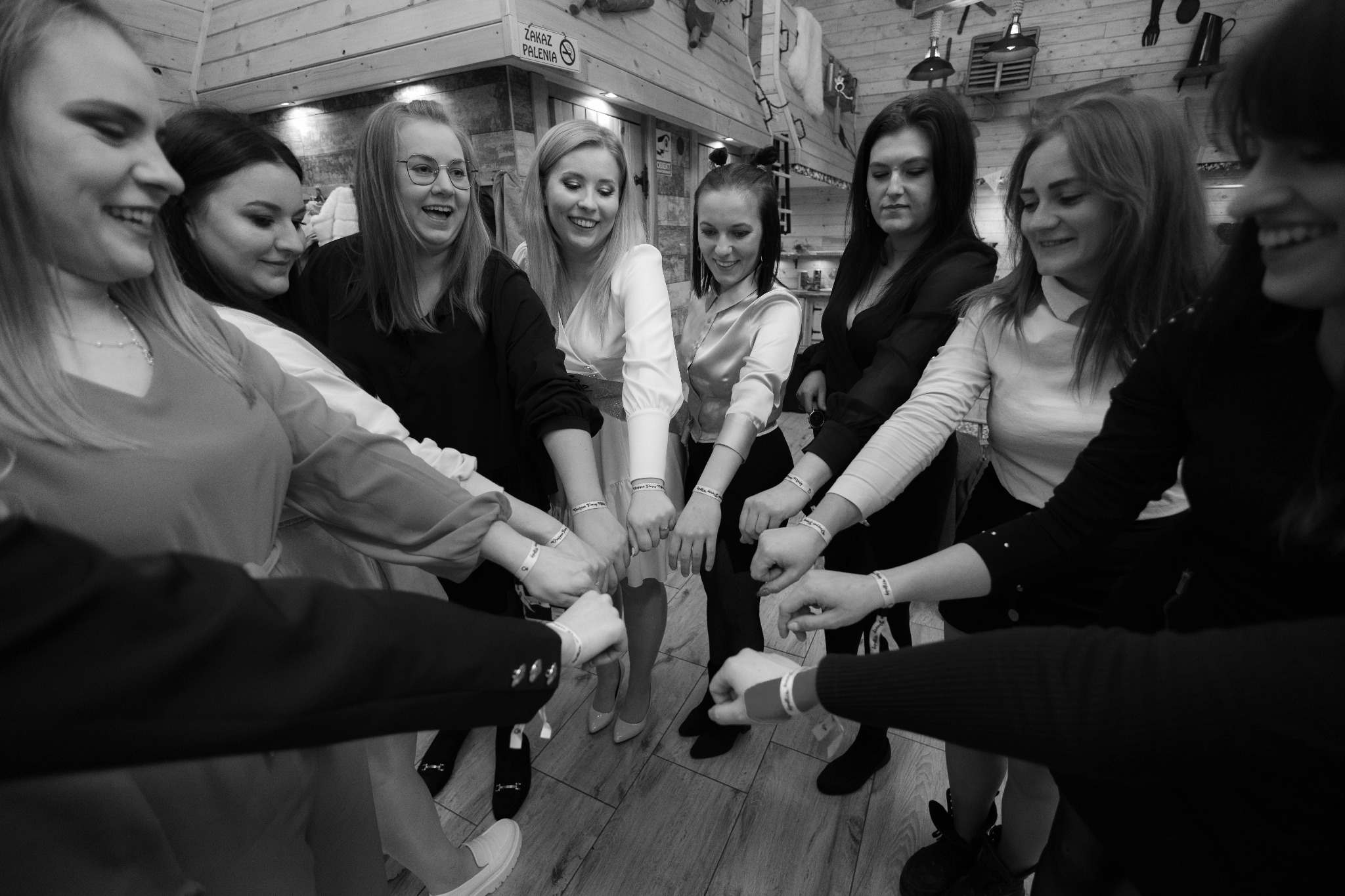 Czarno-białe zdjęcie grupy młodych kobiet i mężczyzn, stojących w kręgu i trzymających się za ręce, z widocznymi identyfikatorami na nadgarstkach, w rustykalnym wnętrzu z drewnianymi ścianami...