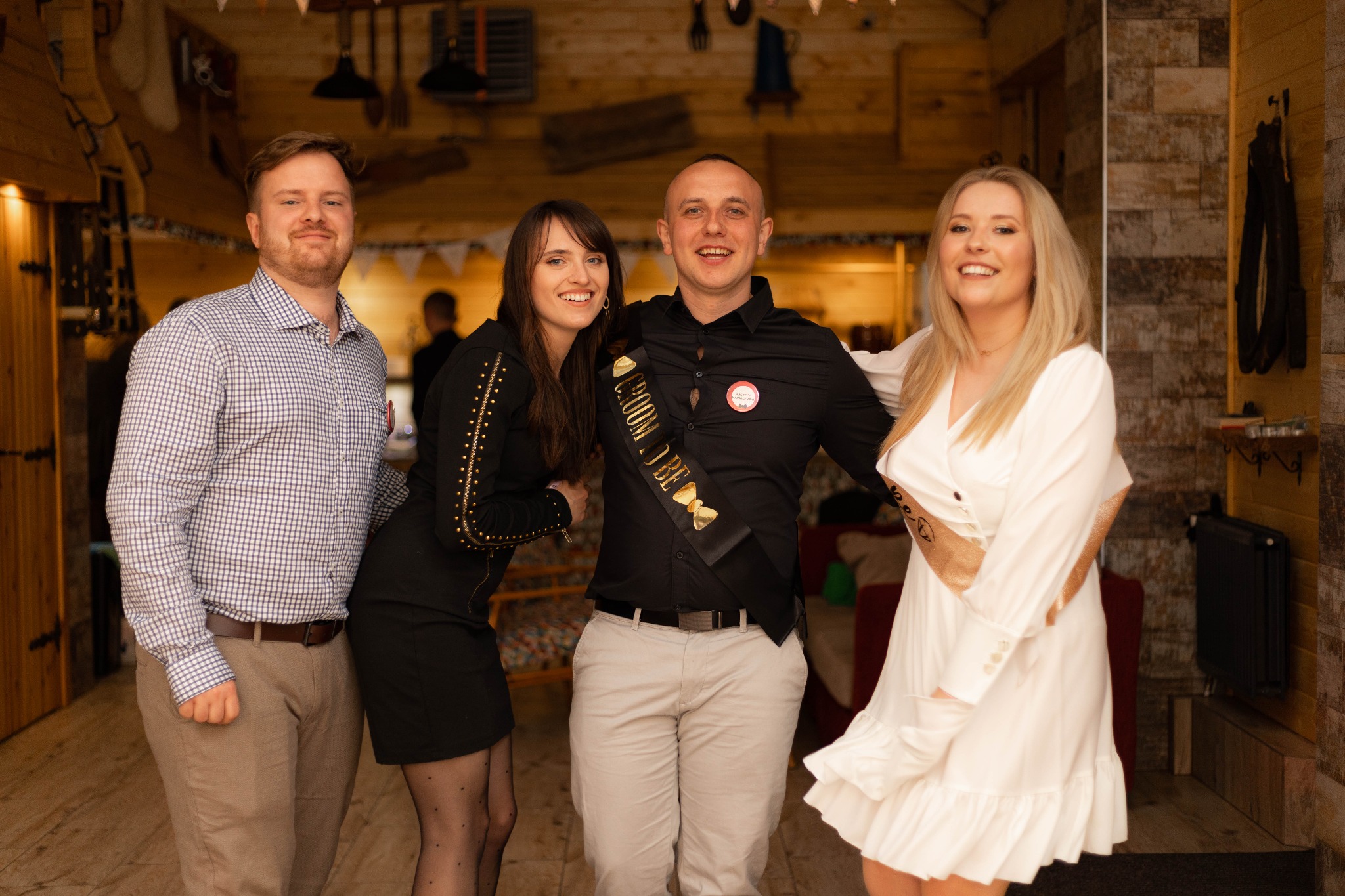 Grupa czterech uśmiechniętych osób pozuje do zdjęcia w drewnianym wnętrzu; mężczyzna ma szarfę z napisem 'Groom to be'.