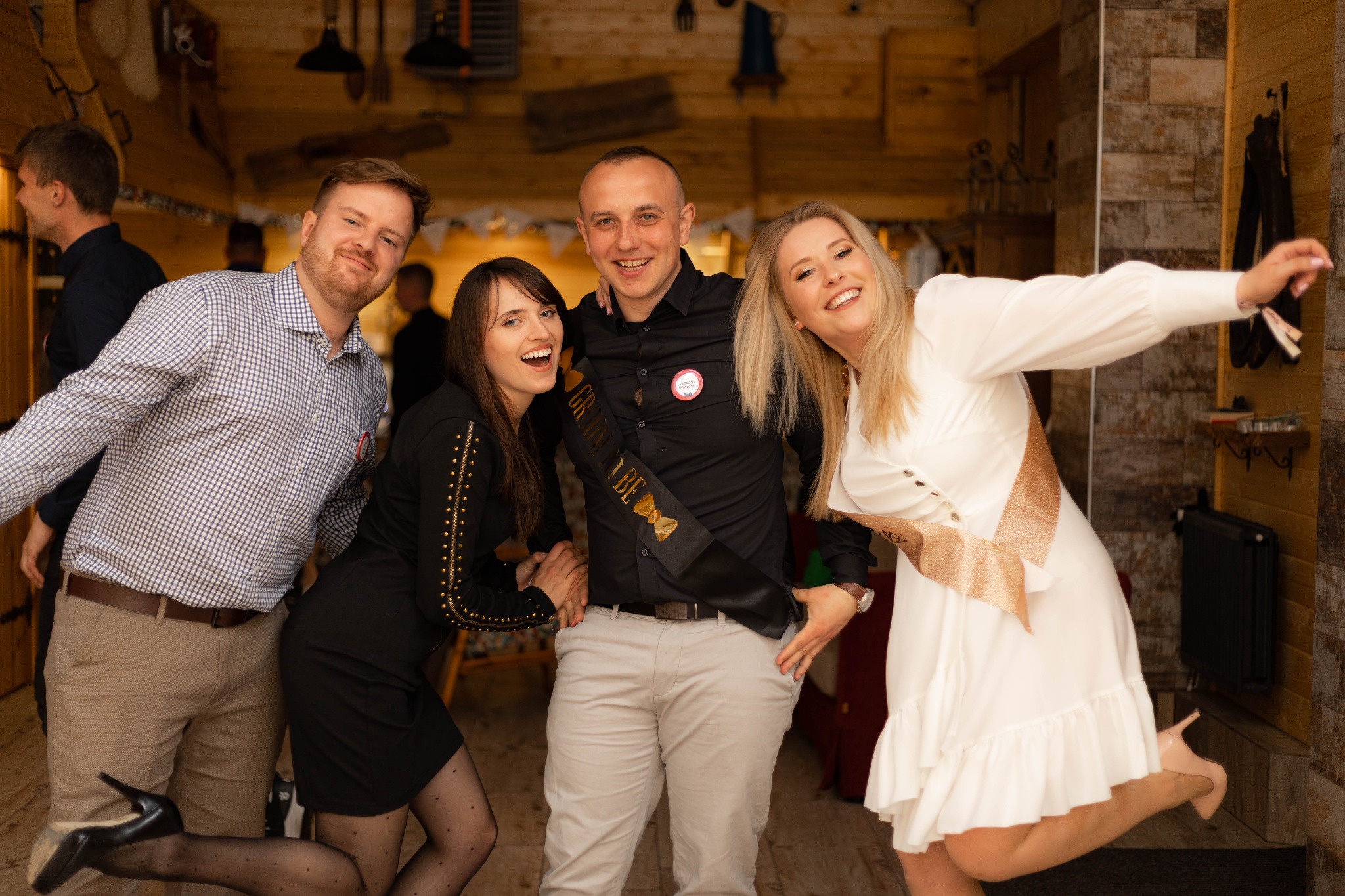 Uśmiechnięta grupa przyjaciół pozuje do zdjęcia w drewnianym wnętrzu podczas wieczoru kawalerskiego, mężczyzna w czarnej koszuli ma szarfę z napisem 'Groom to be'.