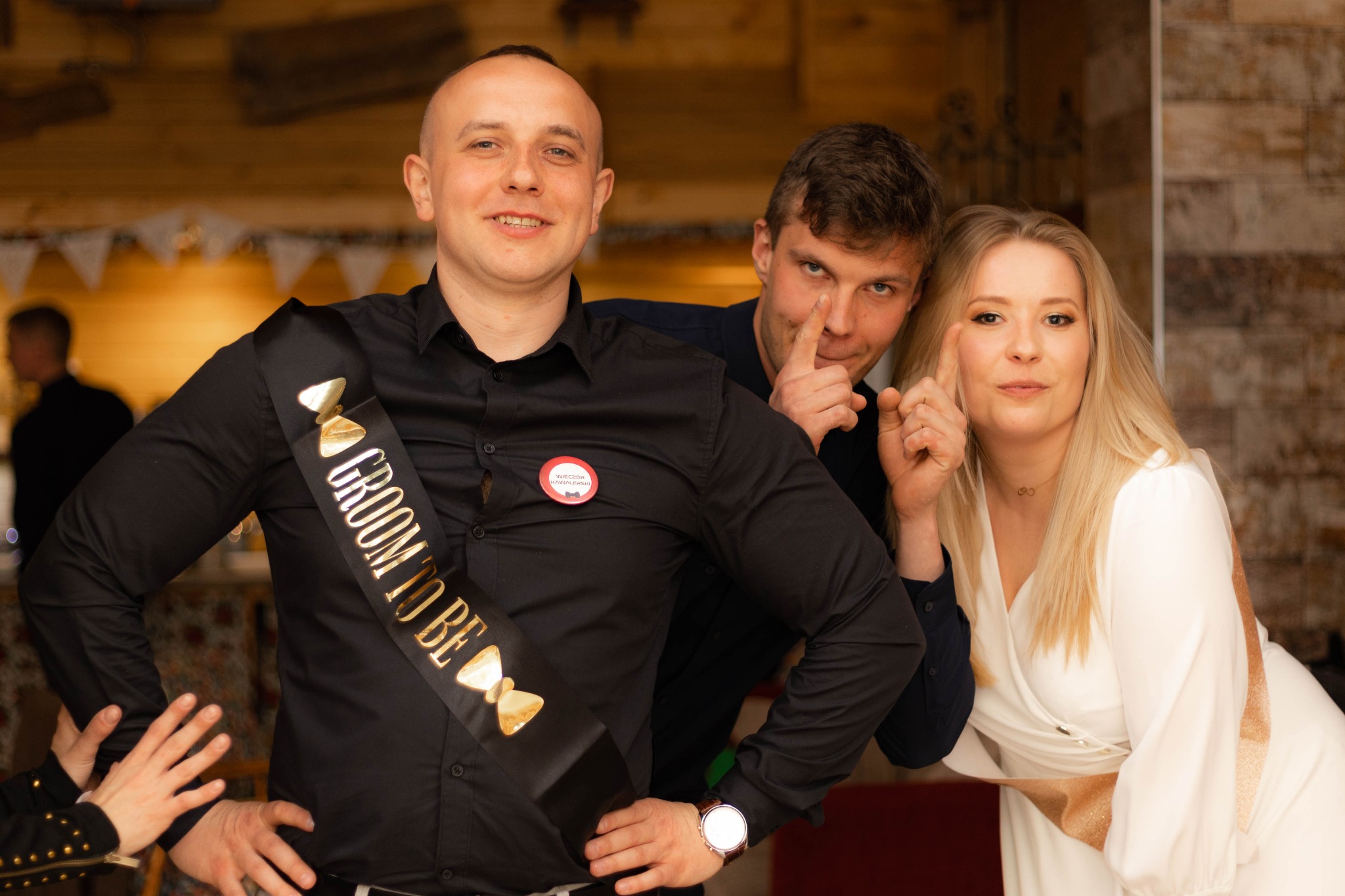 Przyszły pan młody z szarfą 'Groom to be' pozuje z przyjaciółmi podczas wieczoru kawalerskiego, na koszuli przypinka z napisem 'Wieczór Kawalerski'.