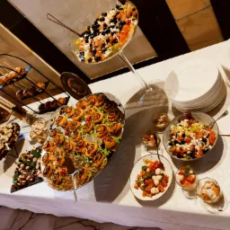 Stół z różnorodnymi przekąskami: roladki z ciasta francuskiego, sałatki w szklanych naczyniach, koreczki, talerze z pomidorami i mozzarellą, oraz sztućce w koszyczkach.