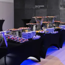 Elegancko zastawiony stół z przekąskami na czarnym obrusie w sali 'Czerwona'. Widoczne różnorodne przystawki, talerze i dekoracyjne stojaki, oświetlone niebieskim światłem.