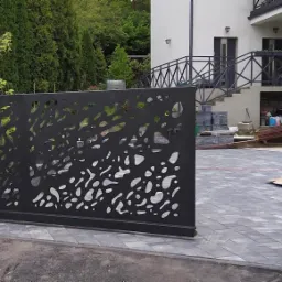 Przesuwna brama wjazdowa z czarnego metalu, o ażurowym wzorze z organicznymi kształtami, osadzona między kamiennymi słupkami na tle domu z zewnętrznymi schodami.