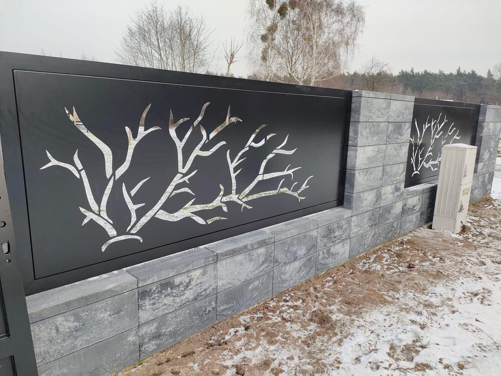 Nowoczesne ogrodzenie z czarnych paneli z wycinanym laserowo motywem drzewa, osadzone na szarych, betonowych słupkach i podmurówce, częściowo pokryte śniegiem.