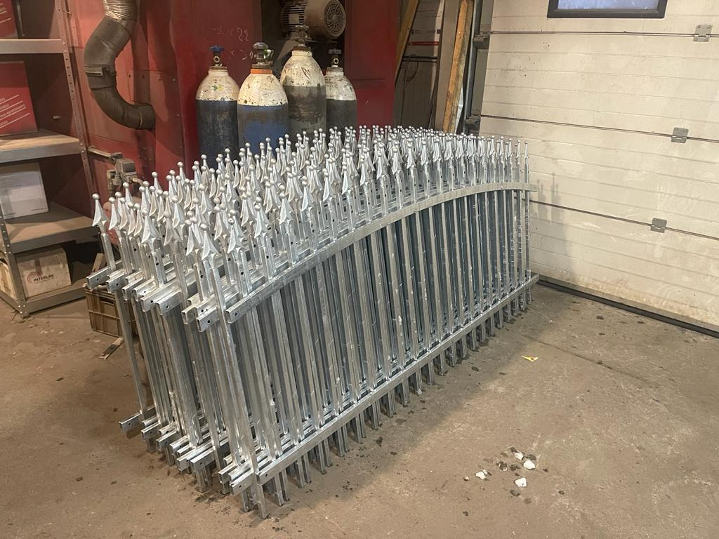 Stos ocynkowanych elementów ogrodzenia kutego z ozdobnymi grotami, ułożonych w warsztacie na betonowej podłodze obok ściany garażowej i butli z gazem.