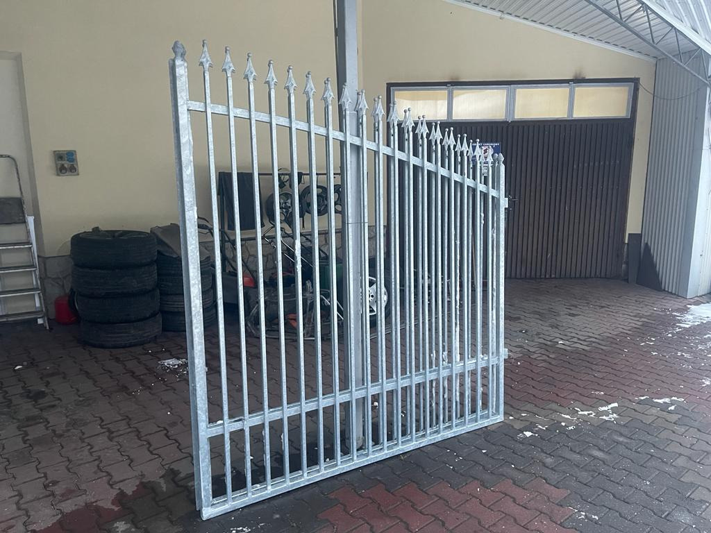 Surowa, metalowa brama wjazdowa z pionowymi prętami i ozdobnymi grotami, przygotowana do montażu w Jarosławiu, widoczna na tle garażu i sterty opon.