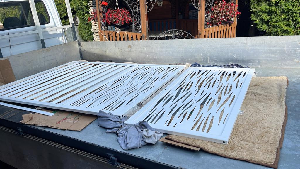 Dwa białe, metalowe panele ogrodzeniowe z abstrakcyjnym wzorem wyciętym laserowo, leżące w skrzyni samochodu dostawczego na kartonie i szmacie, przygotowane do transportu lub montażu.