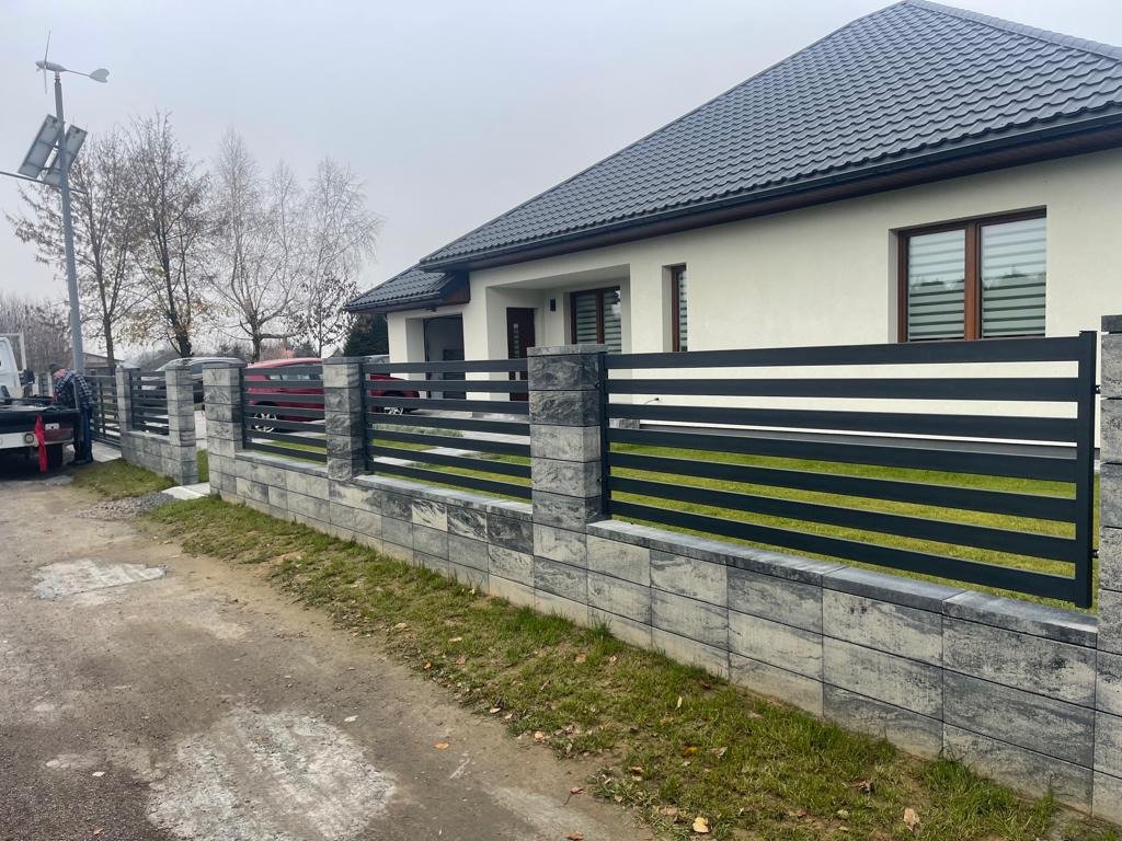 Nowoczesne ogrodzenie metalowe z poziomymi panelami w kolorze czarnym i białym, zamontowane na murku z szarej kostki, na tle białego domu z ciemną dachówką i pochmurnego nieba.