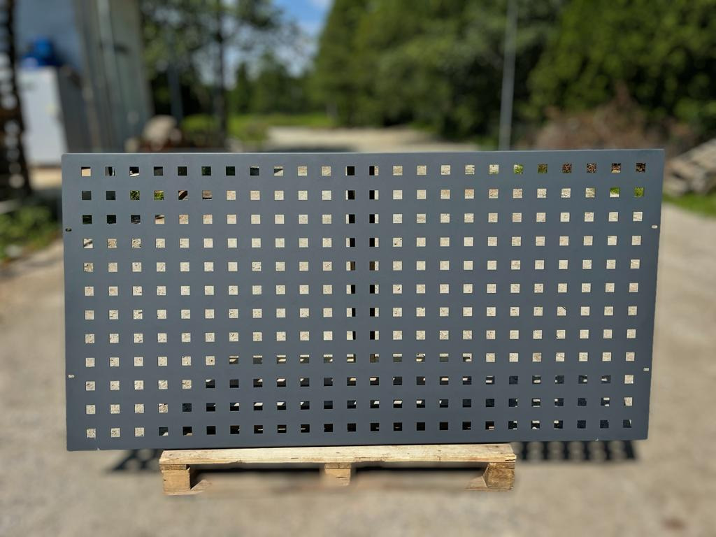 Szary, metalowy panel z regularnymi kwadratowymi otworami, umieszczony na drewnianej palecie na zewnątrz.