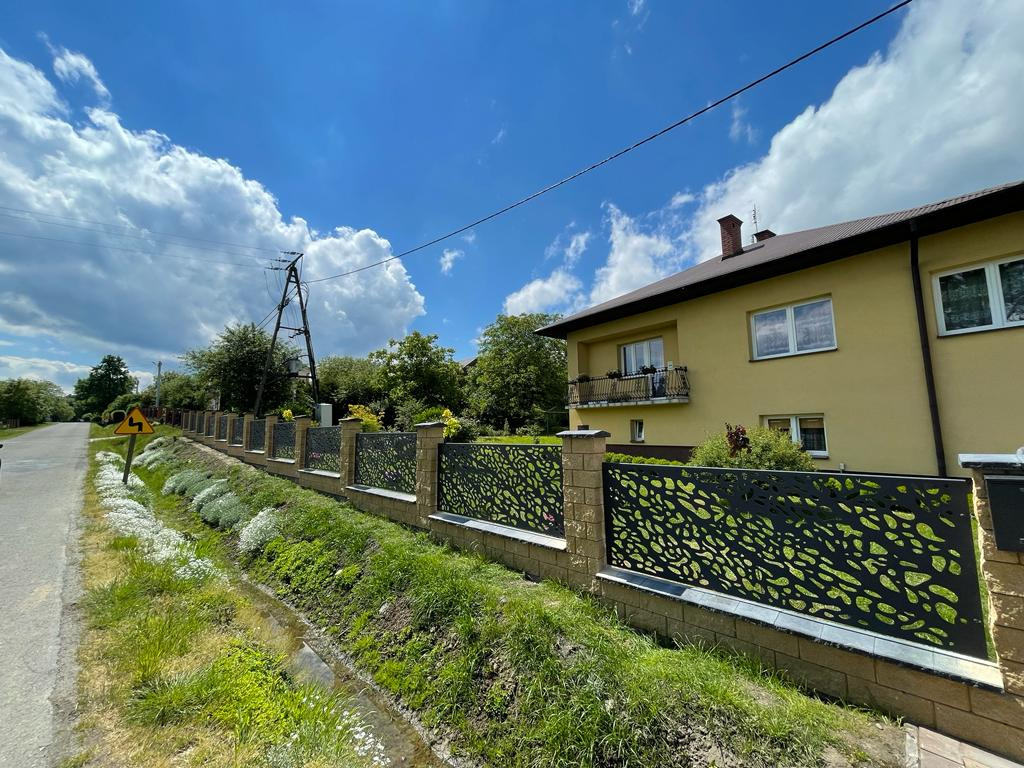 Nowoczesne ogrodzenie z ażurowymi, metalowymi panelami na niskiej podmurówce z kostki, otoczone zielenią i kwiatami, na tle domu z balkonem i błękitnego nieba.