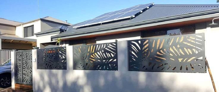 Nowoczesne ogrodzenie z metalowych paneli z wycinanym wzorem liści, zamontowane na niskim murku, w tle dom z panelami słonecznymi na dachu.