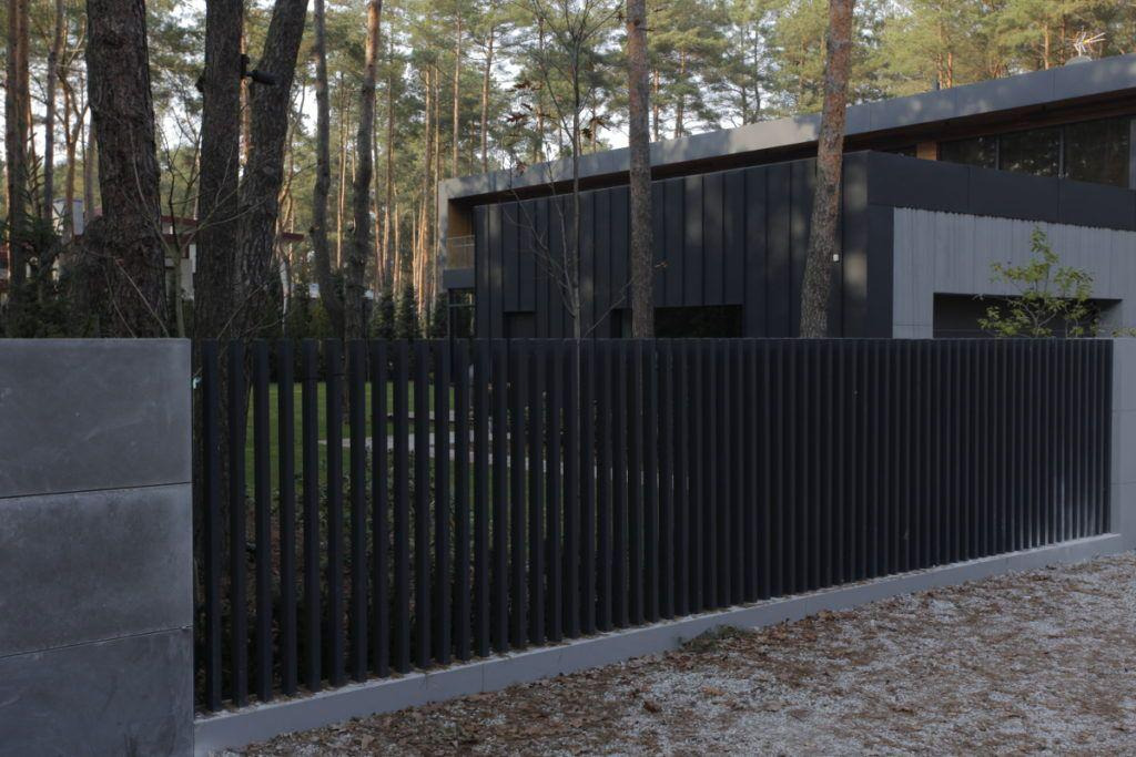 Nowoczesne ogrodzenie z pionowych, czarnych profili metalowych, z szarymi słupkami z betonu architektonicznego, na tle domu w leśnej scenerii.