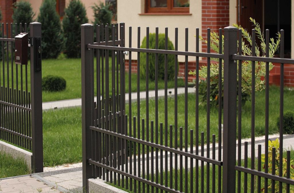 Nowoczesne ogrodzenie z metalowych prętów w kolorze grafitowym, z widoczną skrzynką pocztową i fragmentem domu w tle.