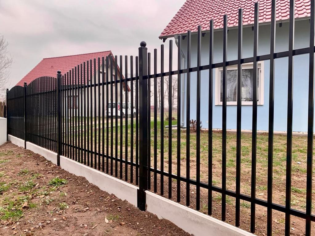 Czarne, pionowe ogrodzenie z metalowych prętów z betonową podmurówką, na tle niebieskiego domu z czerwoną dachówką w pochmurny dzień.