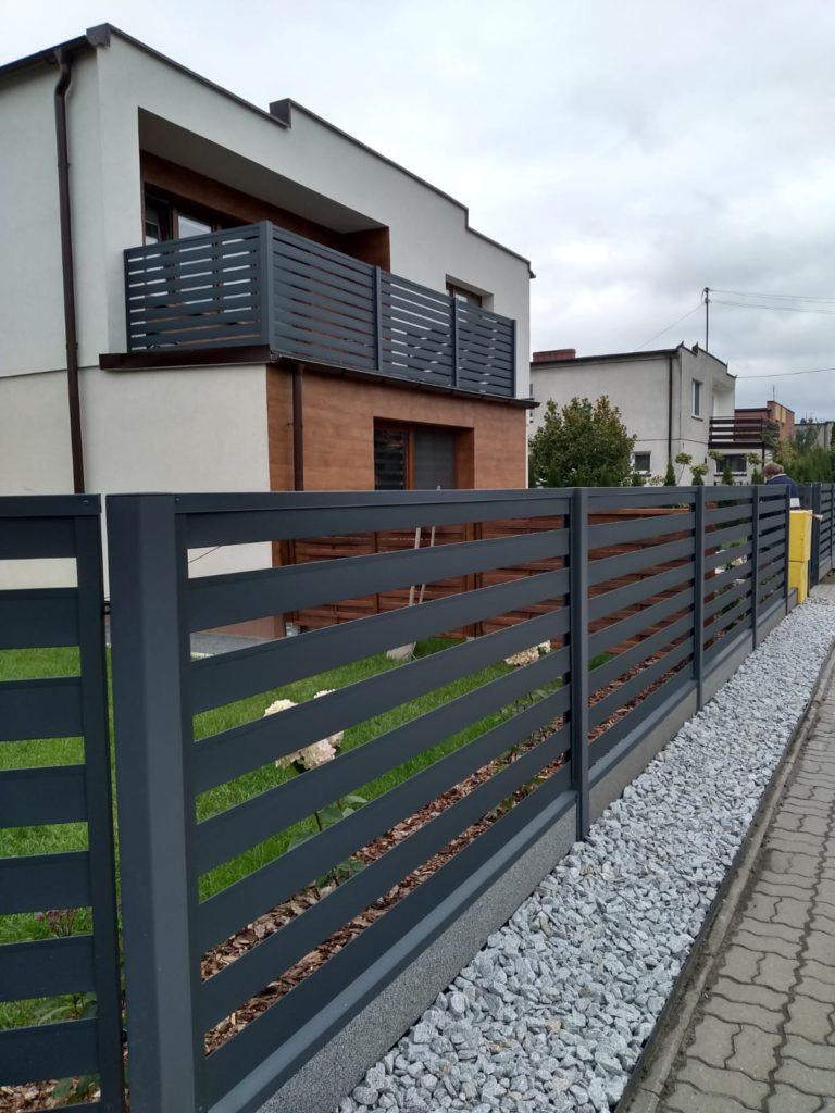 Nowoczesne ogrodzenie z poziomych, szarych profili, widok z zewnątrz na tle domu z balkonem o podobnej stylistyce, wykończenie z białego grysu.