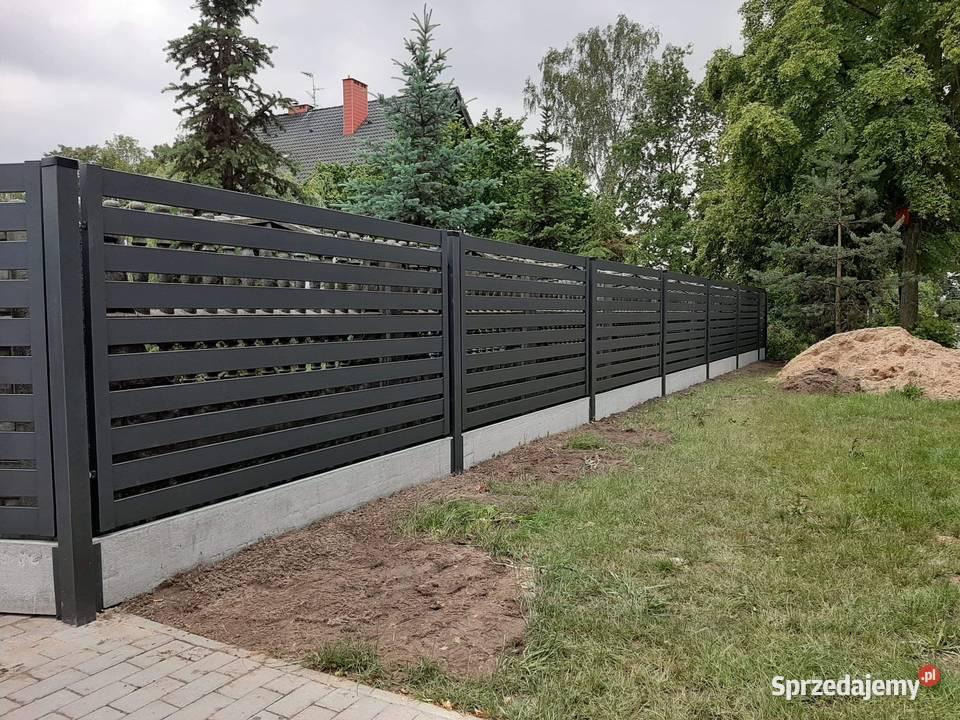 Nowoczesne, poziome ogrodzenie z czarnych profili metalowych na betonowej podmurówce, widok z zewnątrz na tle zieleni i domu z czerwonym dachem.