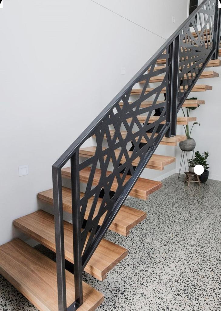 Nowoczesna balustrada schodowa z metalu o geometrycznym wzorze, drewniane stopnie, szary lastryko na podłodze, minimalistyczne wnętrze z roślinami doniczkowymi w tle.