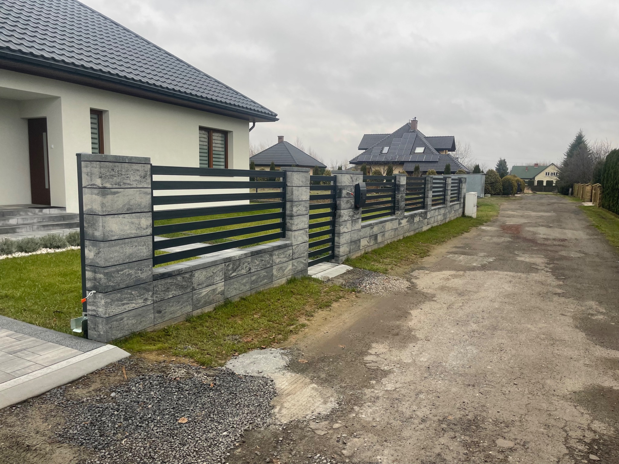 Nowoczesne ogrodzenie panelowe z elementami betonowymi i metalowymi przęsłami w kolorze antracytowym, widoczne na tle domów jednorodzinnych w pochmurny dzień.