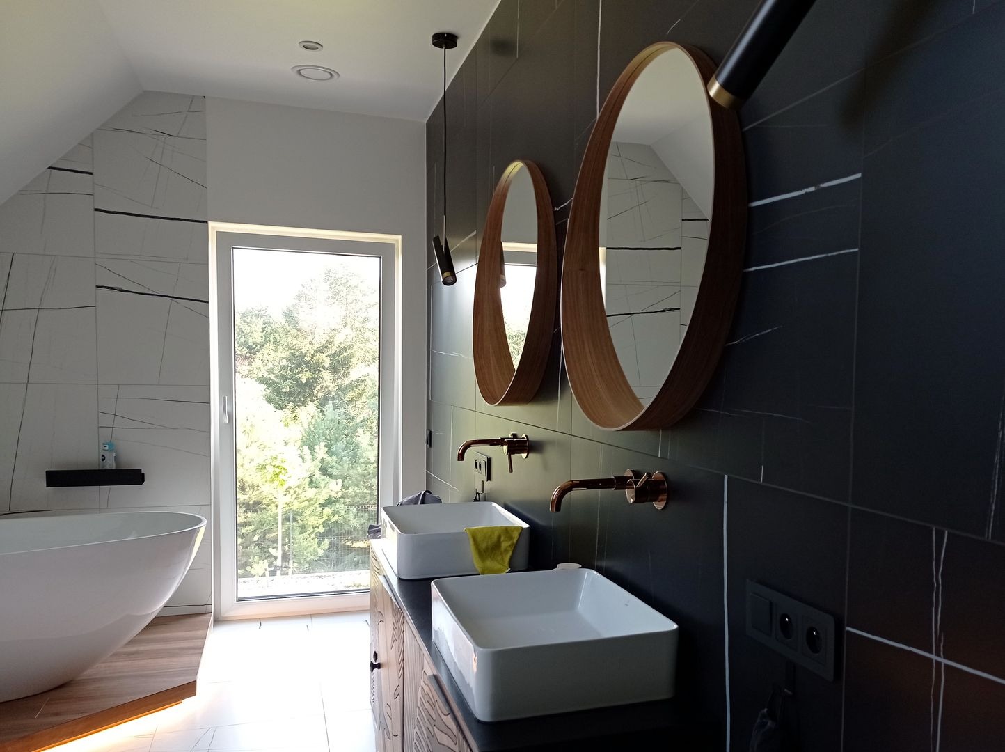 Nowoczesna łazienka na poddaszu z czarnymi płytkami imitującymi marmur, dwoma okrągłymi lustrami w drewnianych ramach, białymi umywalkami nablatowymi i miedzianą armaturą.