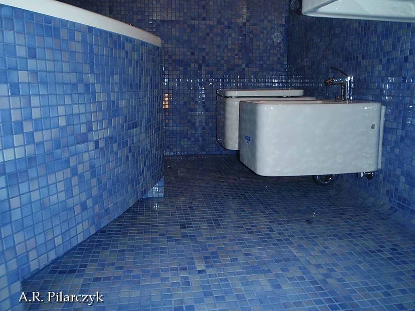 Łazienka wykończona mozaiką w odcieniach błękitu, z dwoma podwieszanymi bidetami i chromowaną armaturą.