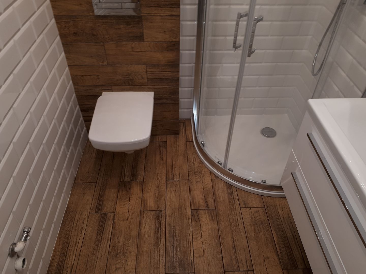 Nowoczesna łazienka z brązowymi płytkami imitującymi drewno na podłodze i ścianie za toaletą, białe płytki na pozostałych ścianach, kabina prysznicowa z przezroczystego szkła i biała szafka...