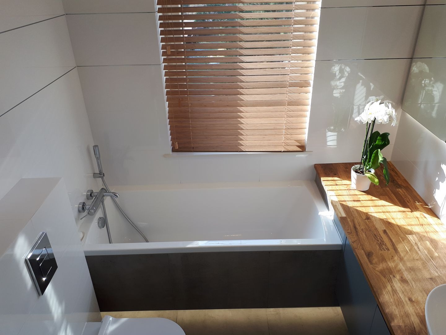 Jasna łazienka z białą wanną obudowaną szarą płytką, drewnianym blatem pod oknem z żaluzjami, białą orchideą w doniczce i chromowaną armaturą.