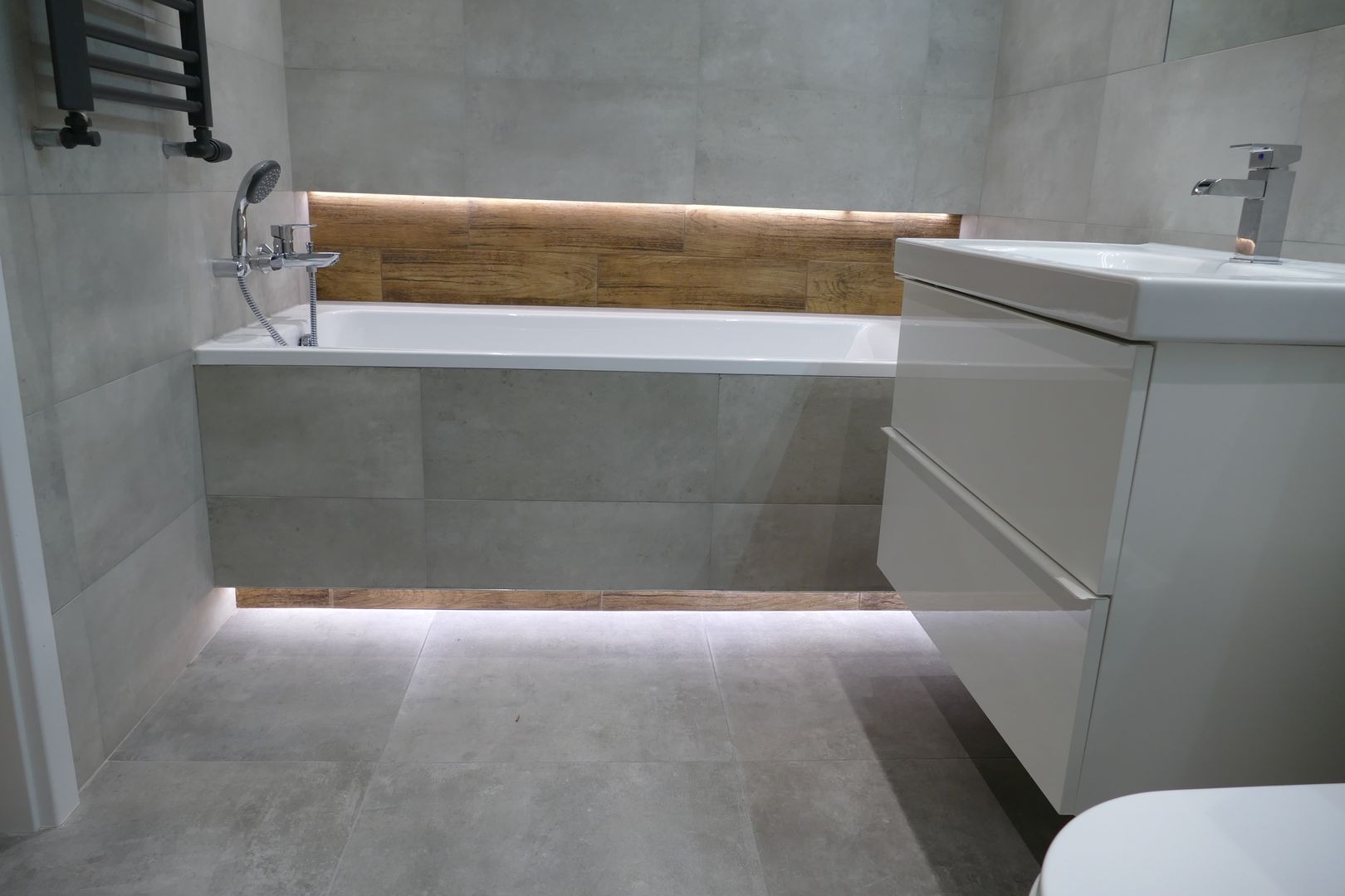 Nowoczesna łazienka z szarymi płytkami, podświetlaną wanną i białą szafką umywalkową z dwiema szufladami oraz grzejnikiem drabinkowym.