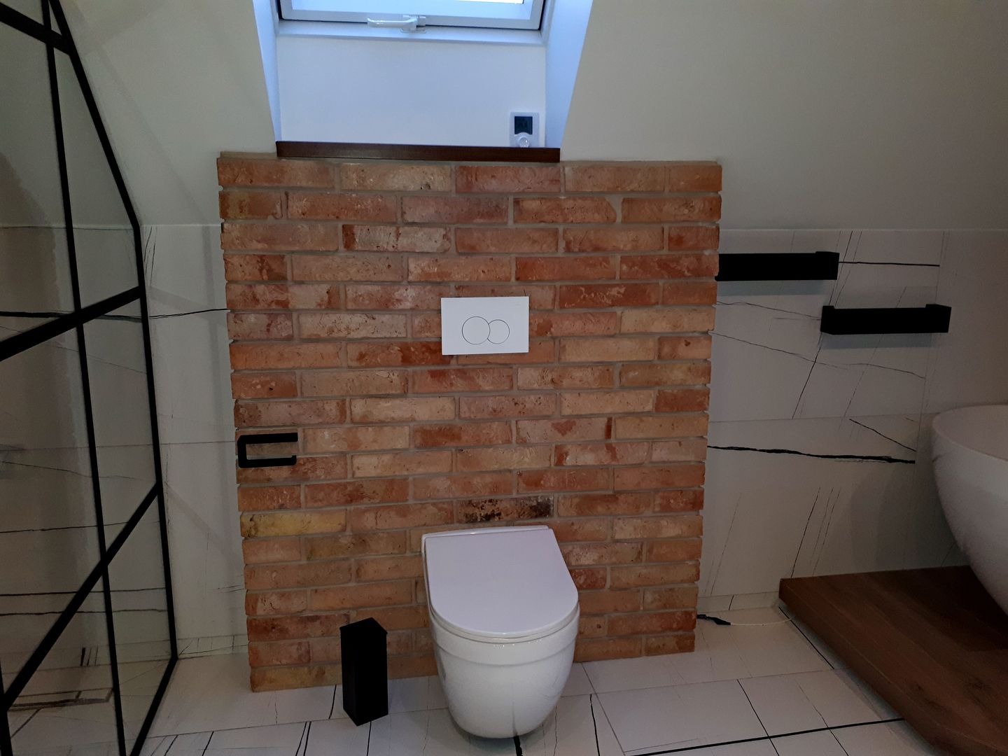 Toaleta w łazience na poddaszu, z obudową ze starej cegły i białymi płytkami imitującymi marmur, czarne półki wiszące i rama kabiny prysznicowej, okno dachowe.