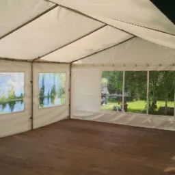 Wnętrze białego namiotu z drewnianą podłogą i oknami z widokiem na zewnątrz, przez które widać zieleń i kwiaty.