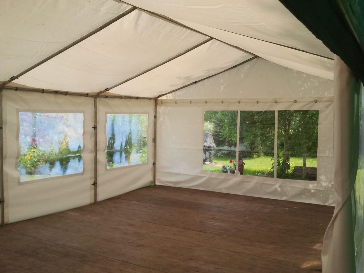 Wnętrze białego namiotu z drewnianą podłogą i oknami z widokiem na zewnątrz, przez które widać zieleń i kwiaty.