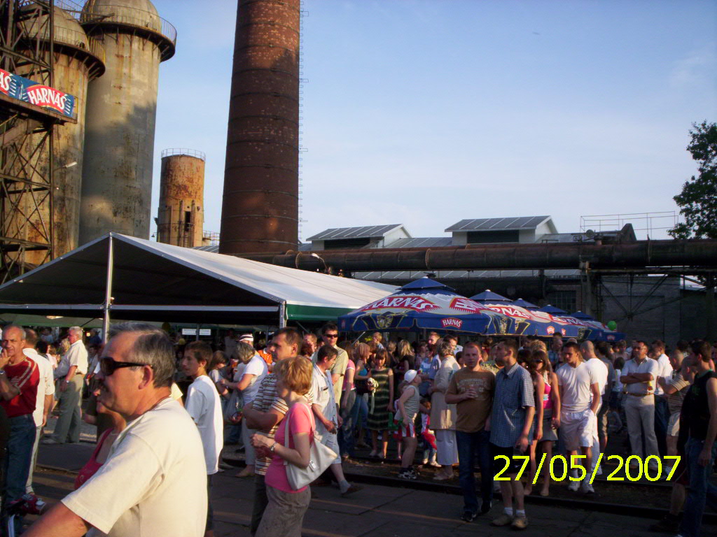 Plenerowa impreza masowa pod białym namiotem i parasolami piwnymi na tle industrialnego krajobrazu z kominami i rurami, tłum ludzi w letnich ubraniach.