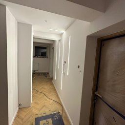 Usługi ogólnobudowlane Tomasz F. - Korytarz po remoncie z parkietem w jodełkę, białymi ścianami z pionowymi lampami LED i zabezpieczonymi drzwiami. Widoczne kable i kartony po płytkach.