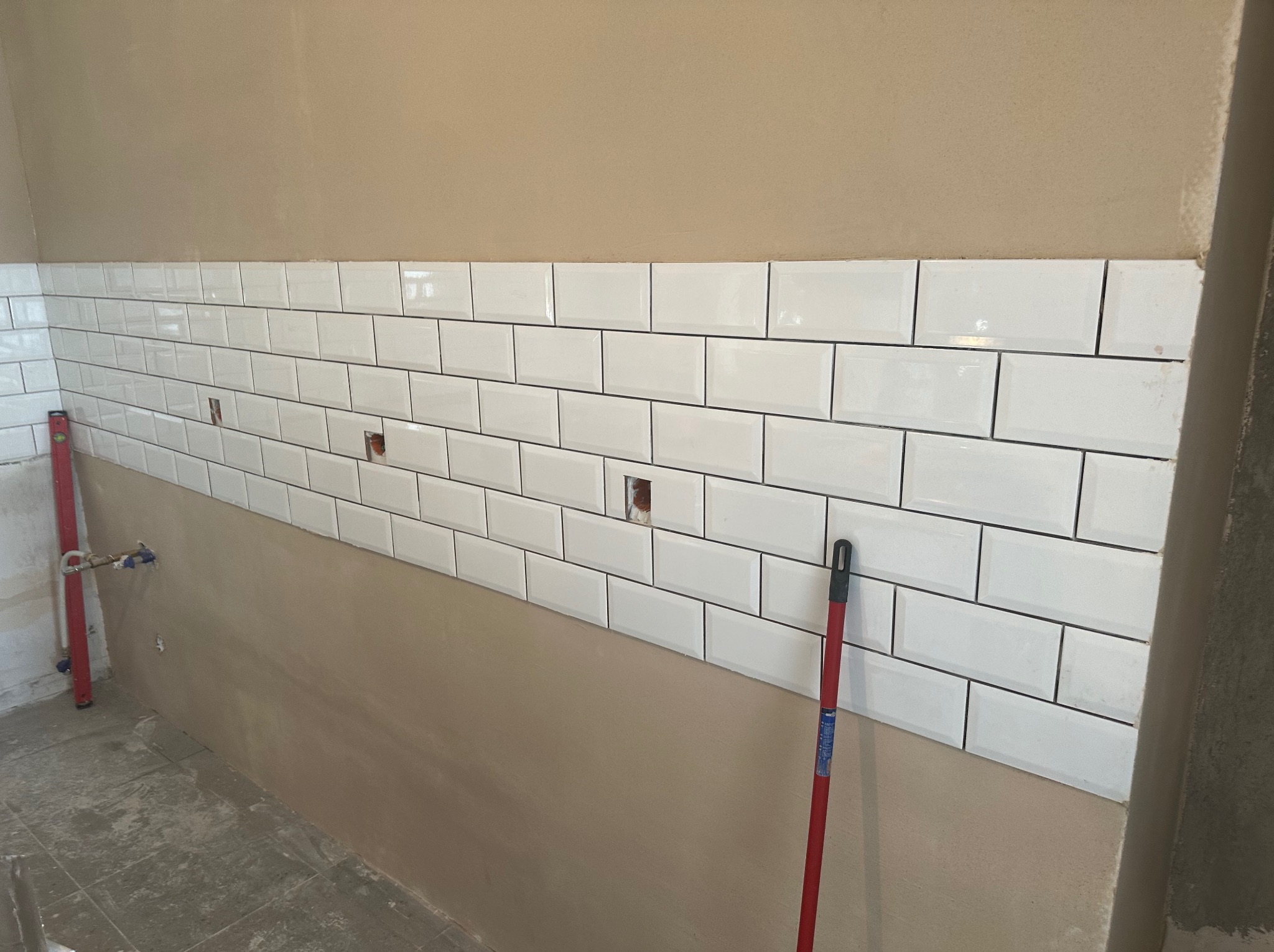 Białe, prostokątne płytki ułożone rzędami na ścianie łazienki, widoczne otwory na instalacje, poziomica oparta o ścianę i mop stojący obok, ściana częściowo przygotowana pod dalsze prace.