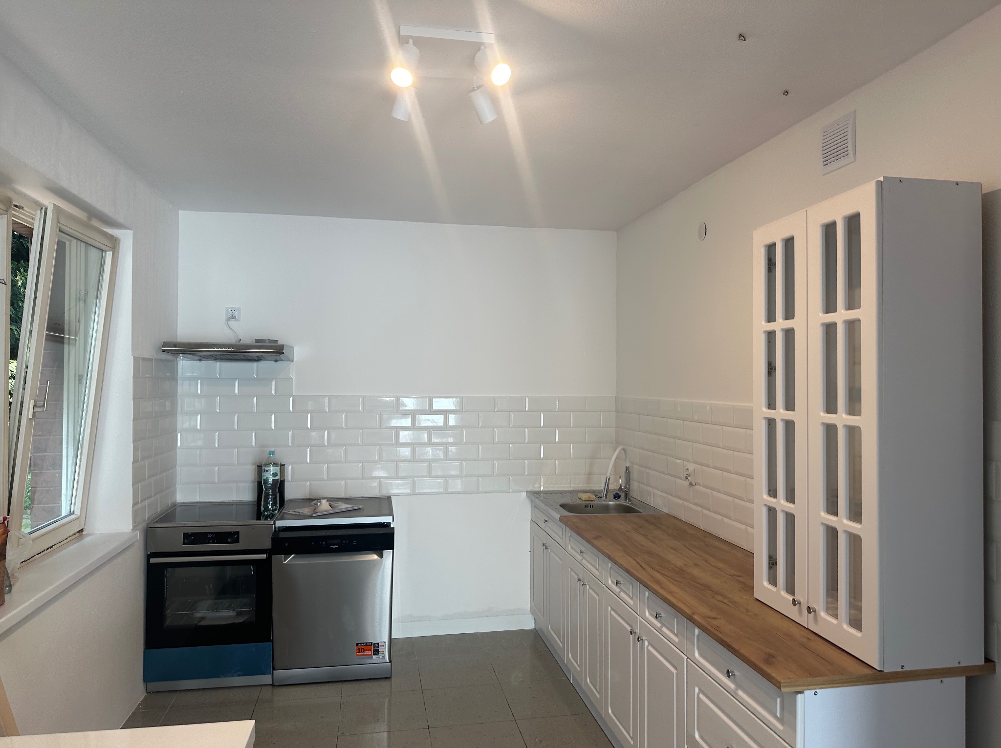 Jasna kuchnia po remoncie z białymi szafkami, drewnianym blatem, białymi płytkami typu metro na ścianie i sprzętem AGD, widok na okno z zielenią na zewnątrz.