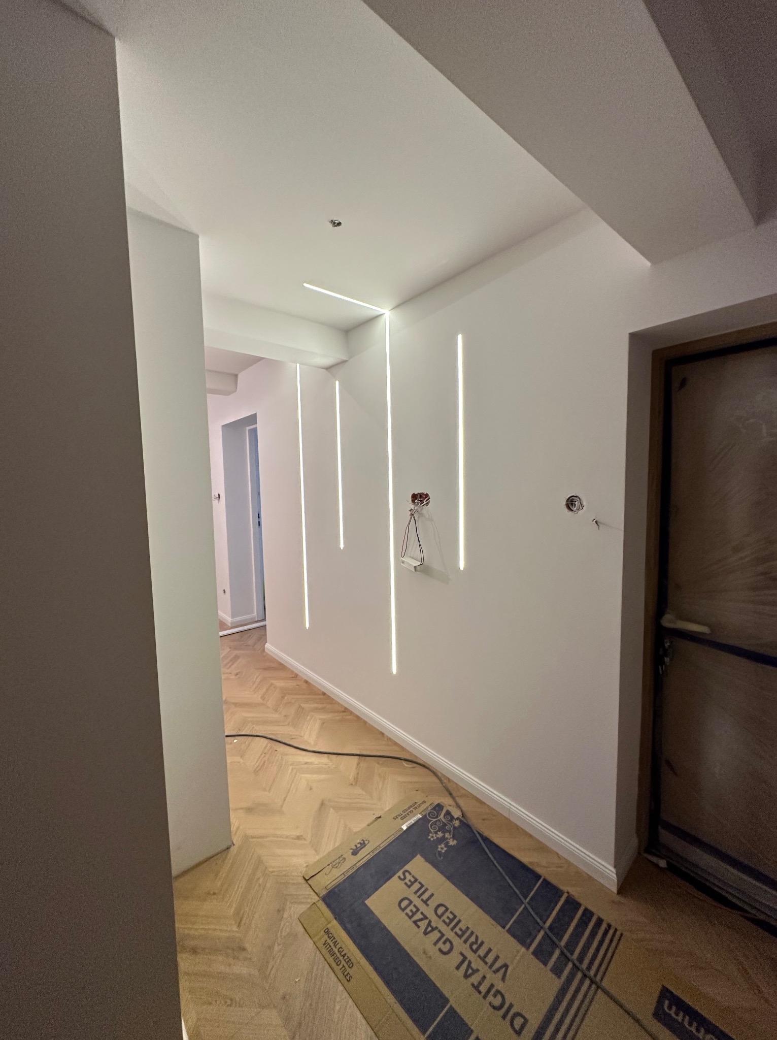 Korytarz po remoncie z podświetlanymi pionowymi liniami LED na ścianach i parkietem ułożonym w jodełkę. Widoczne kartony z płytkami i instalacja elektryczna.