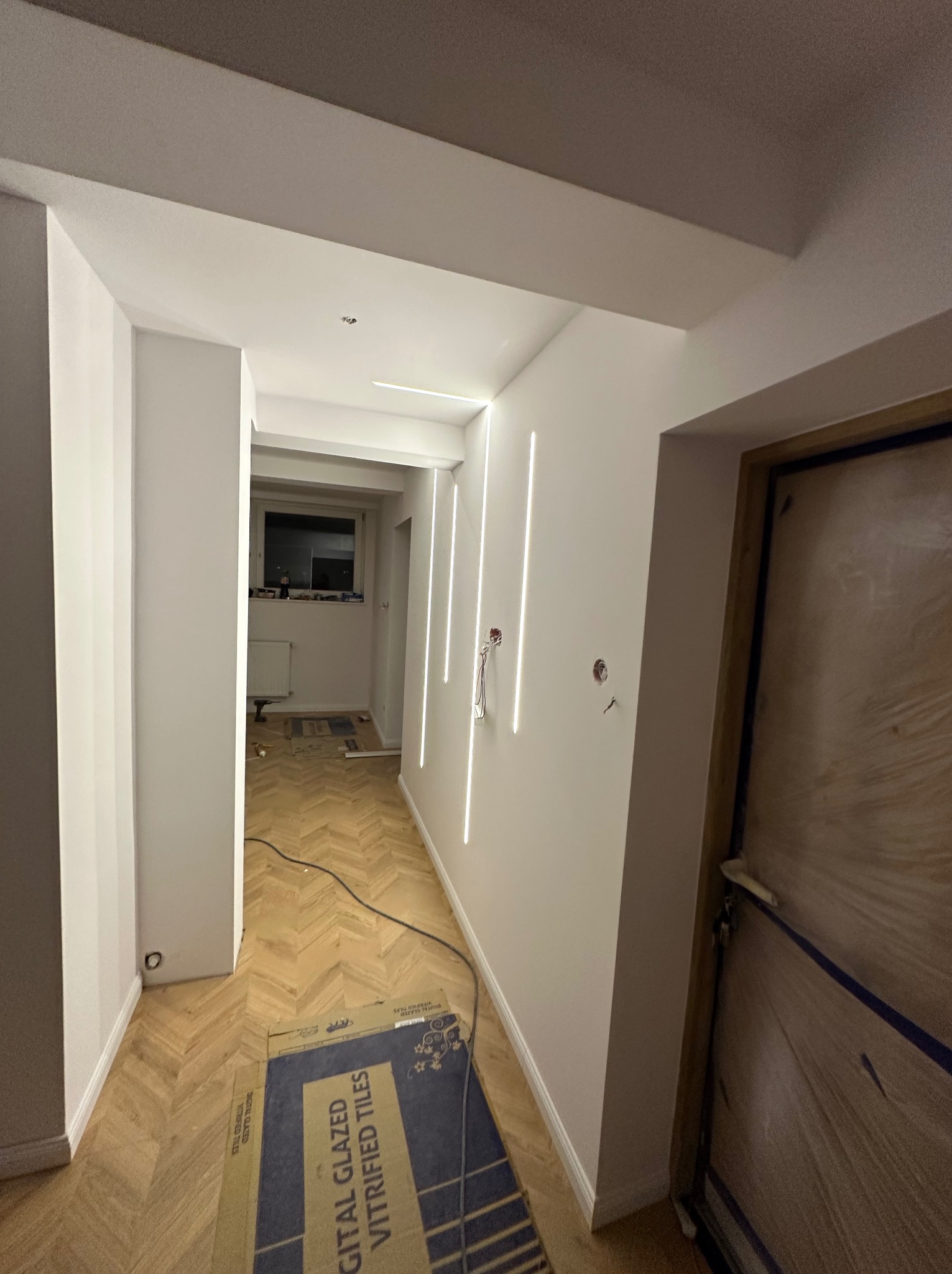 Korytarz po remoncie z parkietem w jodełkę, białymi ścianami z pionowymi lampami LED i zabezpieczonymi drzwiami. Widoczne kable i kartony po płytkach.