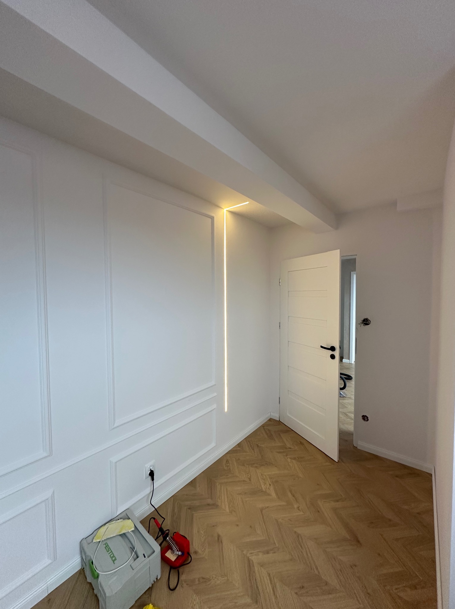 Białe wnętrze po remoncie: ściana z dekoracyjnymi panelami, podłoga w jodełkę, drzwi i pionowa listwa LED. Na podłodze narzędzia.