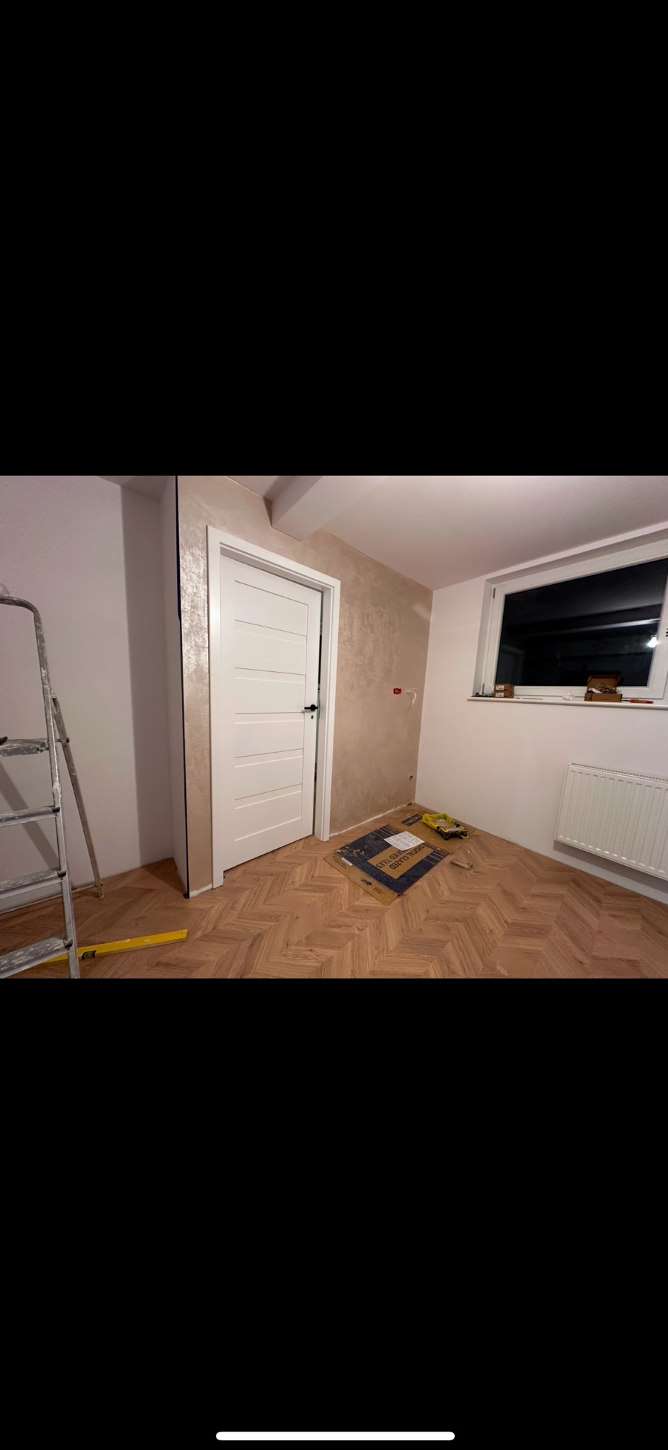 Pokój po remoncie: parkiet w jodełkę, białe drzwi z czarną klamką, pomalowana ściana z widoczną fakturą. U dołu karton z płytkami i narzędzia.