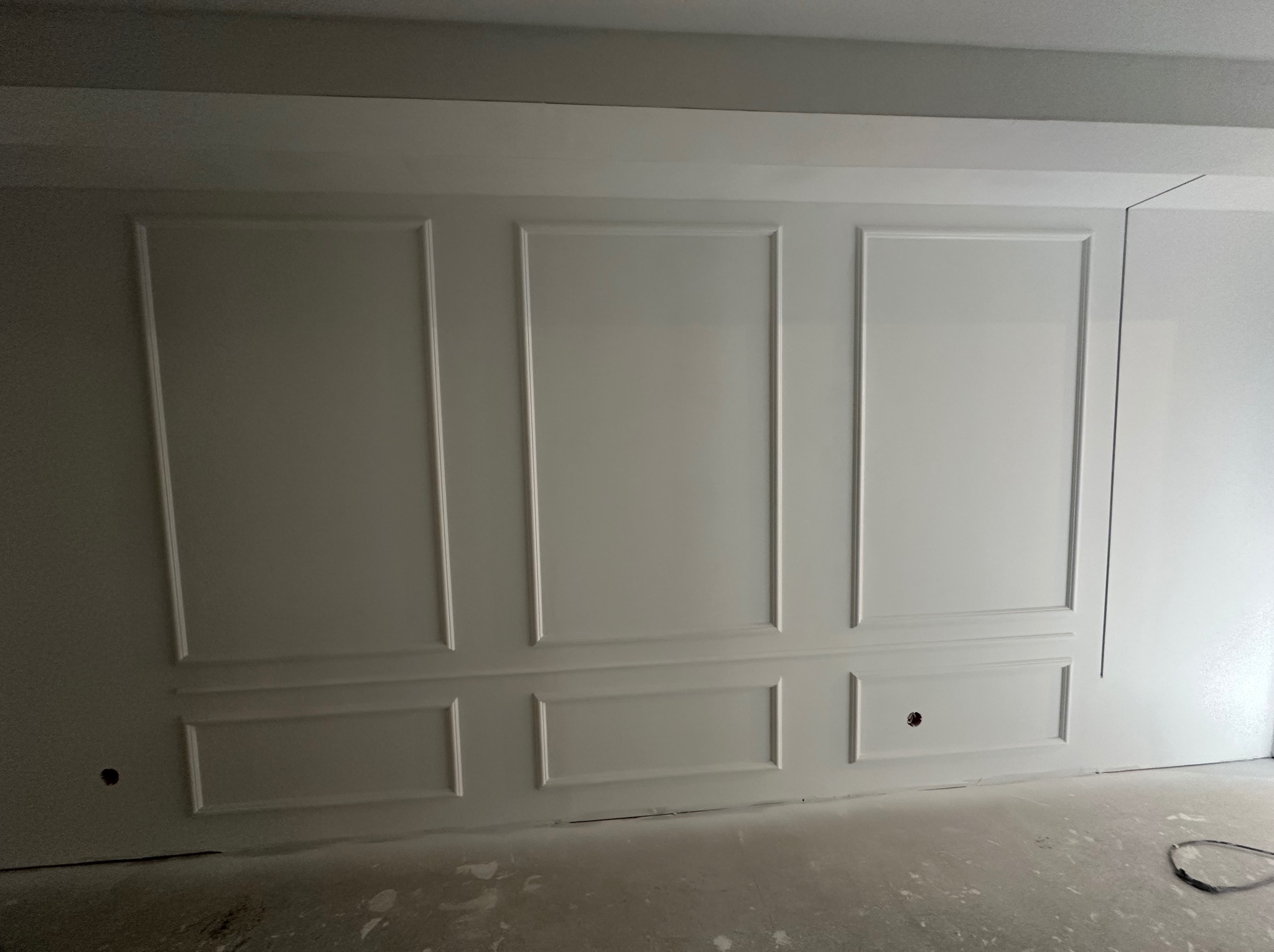 Biała ściana z dekoracyjnymi, symetrycznymi panelami w stylu klasycznym. Widoczne niedoskonałości na podłodze sugerują prace wykończeniowe.