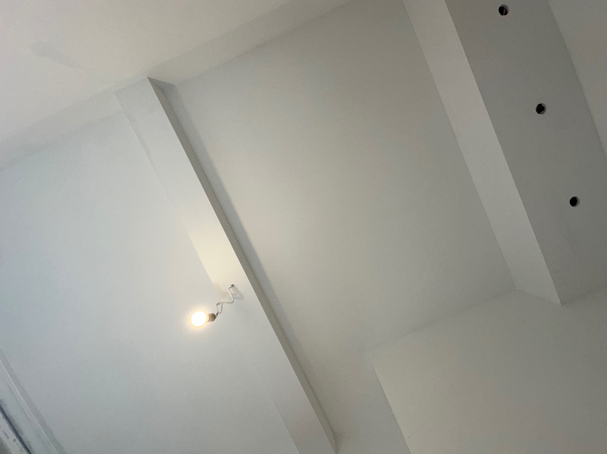 Białe, świeżo pomalowane wnętrze z nieukończonymi punktami oświetleniowymi i widoczną żarówką na kablu, ukazujące etap prac wykończeniowych.