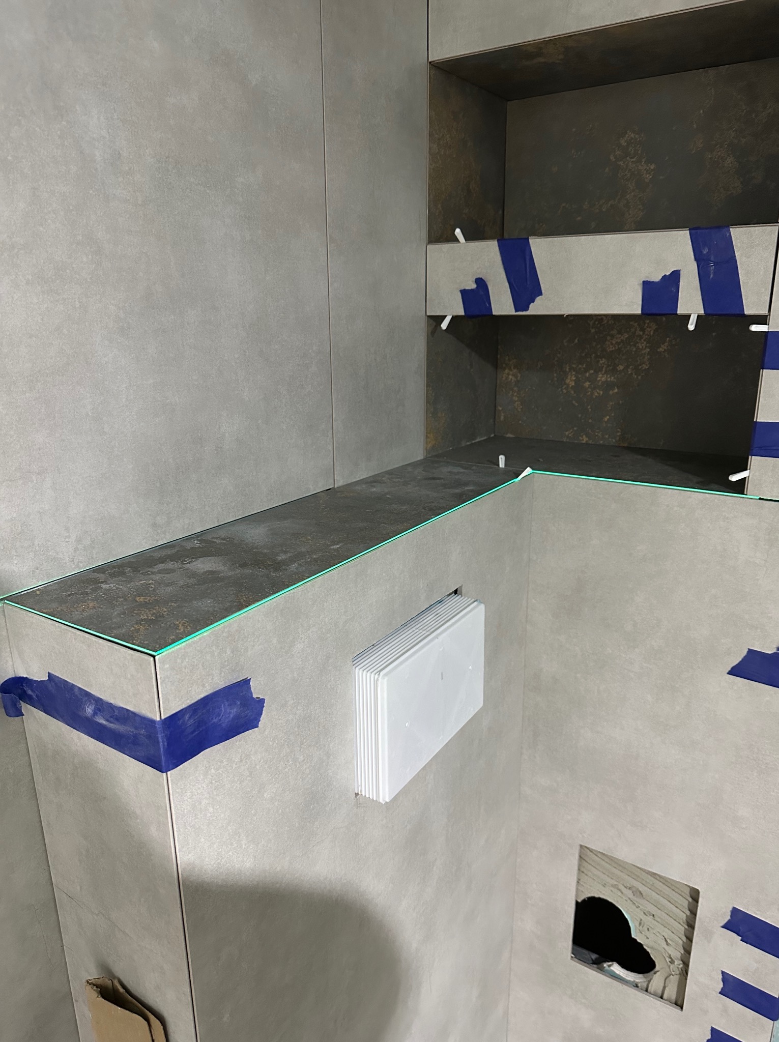 Fragment łazienki w trakcie wykończenia: szare płytki, półki z podświetleniem LED, zabezpieczone taśmą. Widoczny otwór w ścianie i kratka wentylacyjna.