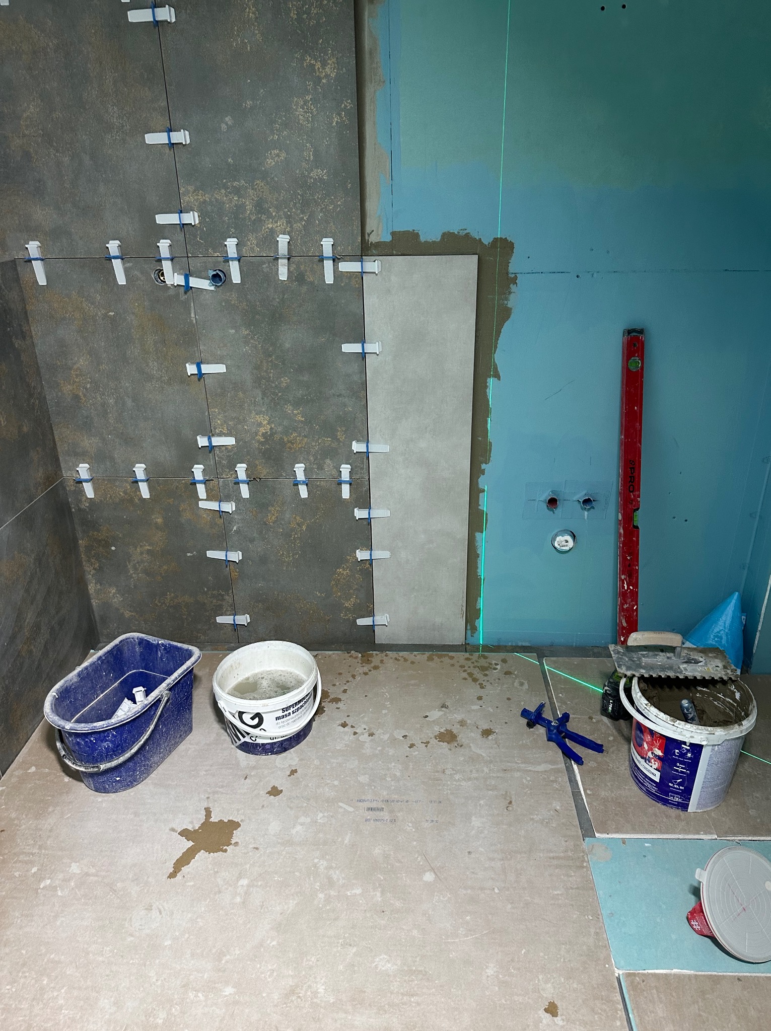 Układanie płytek w łazience: częściowo wykończona ściana z ciemnymi i jasnymi płytkami, poziomica laserowa, wiadra z zaprawą i narzędzia glazurnicze.