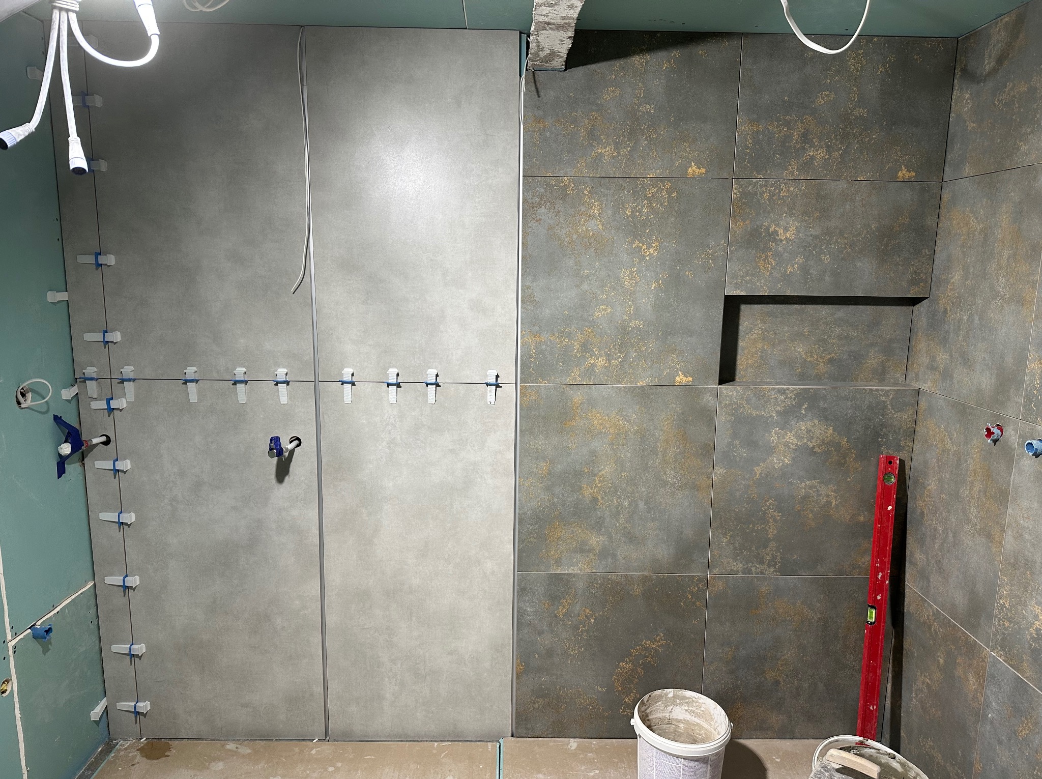 Kładzenie płytek w łazience: szare płytki z fugami i poziomica. Część ściany z płytkami w odcieniach szarości i złota. Widoczne akcesoria montażowe.