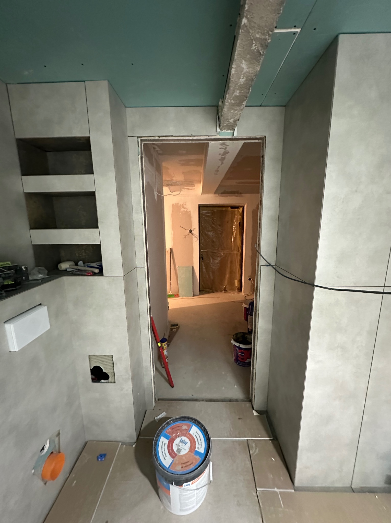Wnętrze łazienki w trakcie wykańczania: szare płytki, otwarta wnęka z półkami, widoczne instalacje i wiadro z klejem. Prace wykończeniowe w toku.