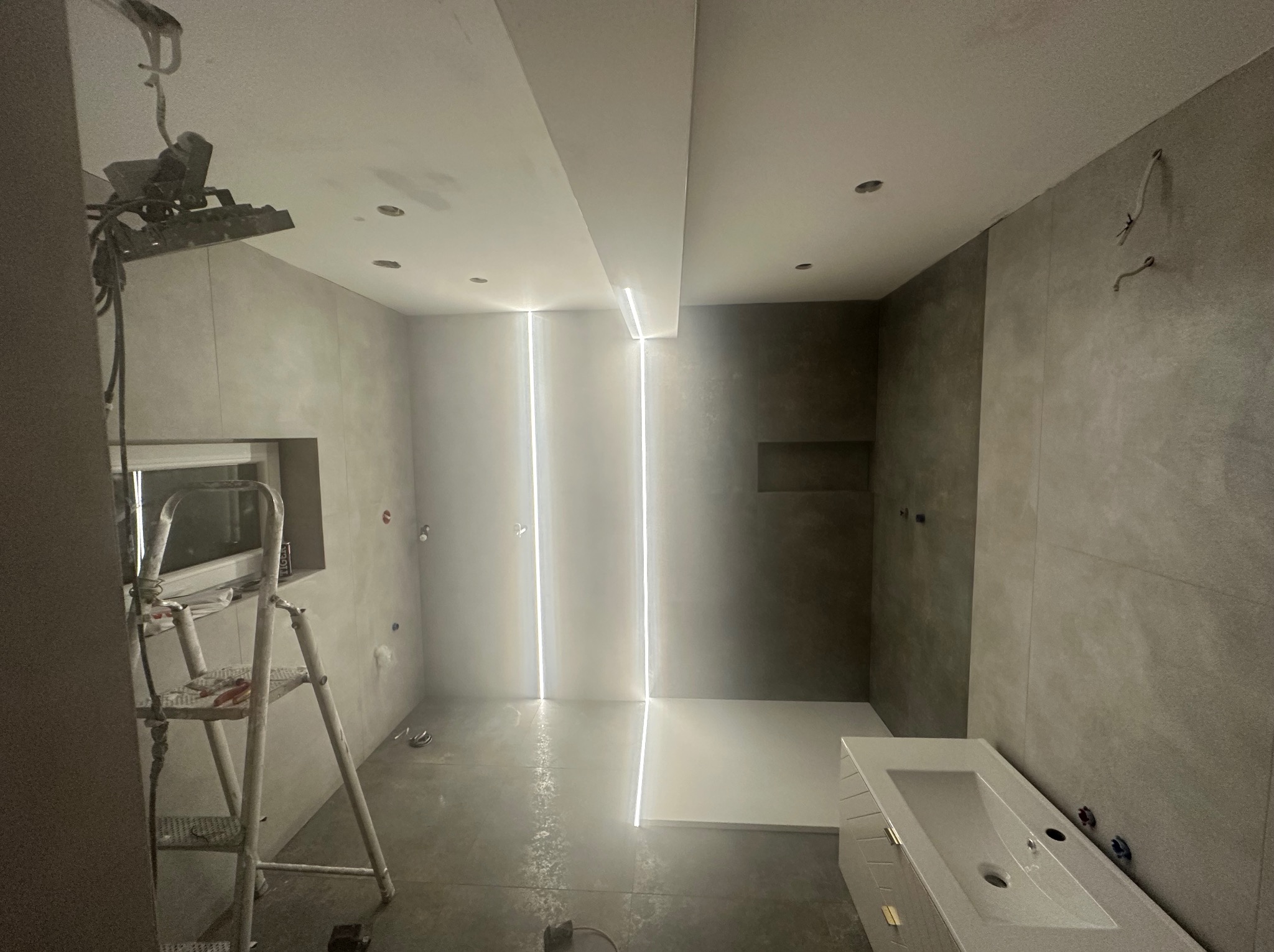 Szara łazienka w trakcie remontu: widoczna drabina malarska, oświetlenie LED i umywalka. Ściany wyłożone płytkami w odcieniach szarości.