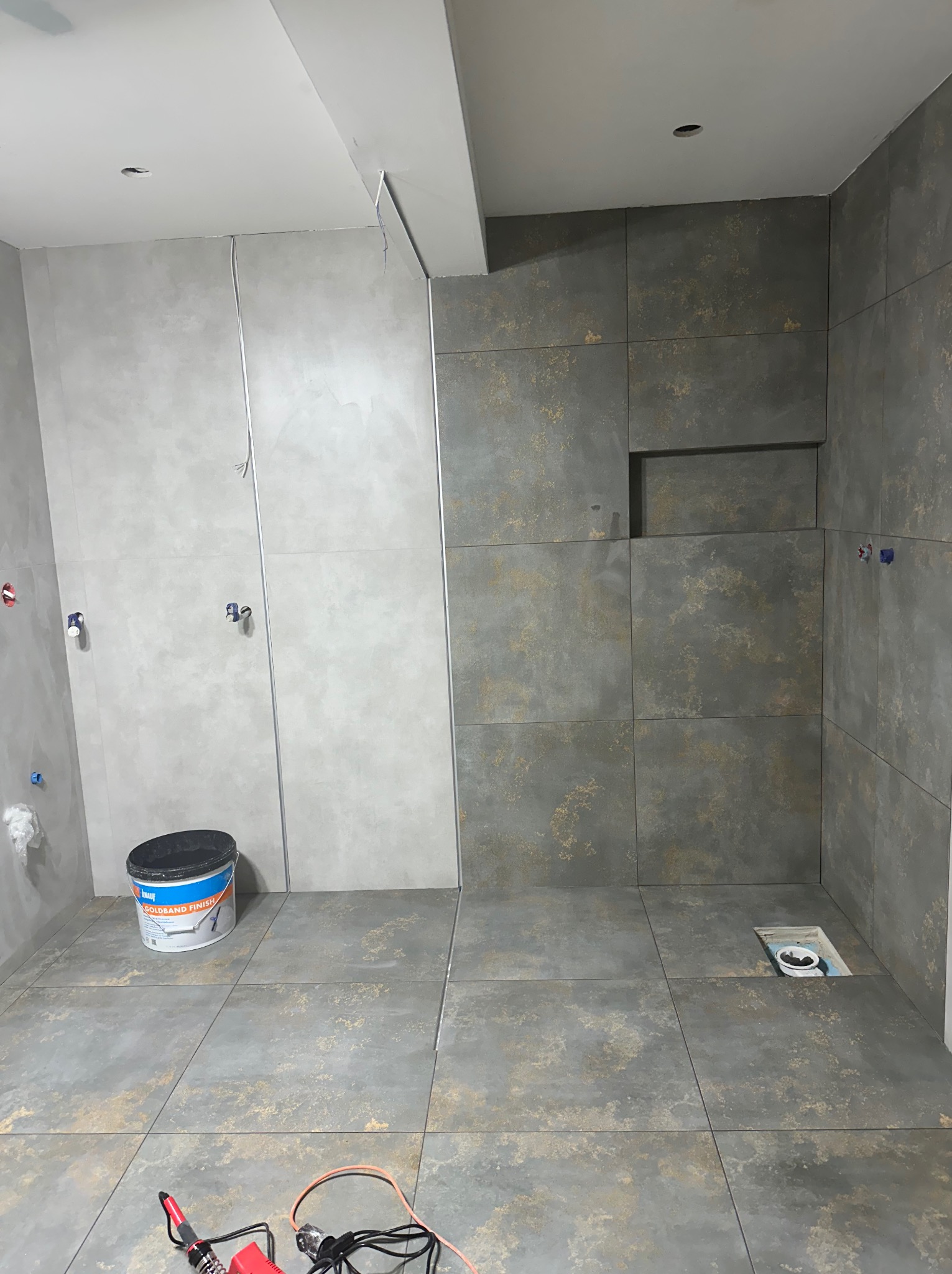 Łazienka w trakcie wykańczania: częściowo ułożone płytki w odcieniach szarości i beżu, widoczne narzędzia, wiadro z zaprawą. Przygotowanie do fugowania.