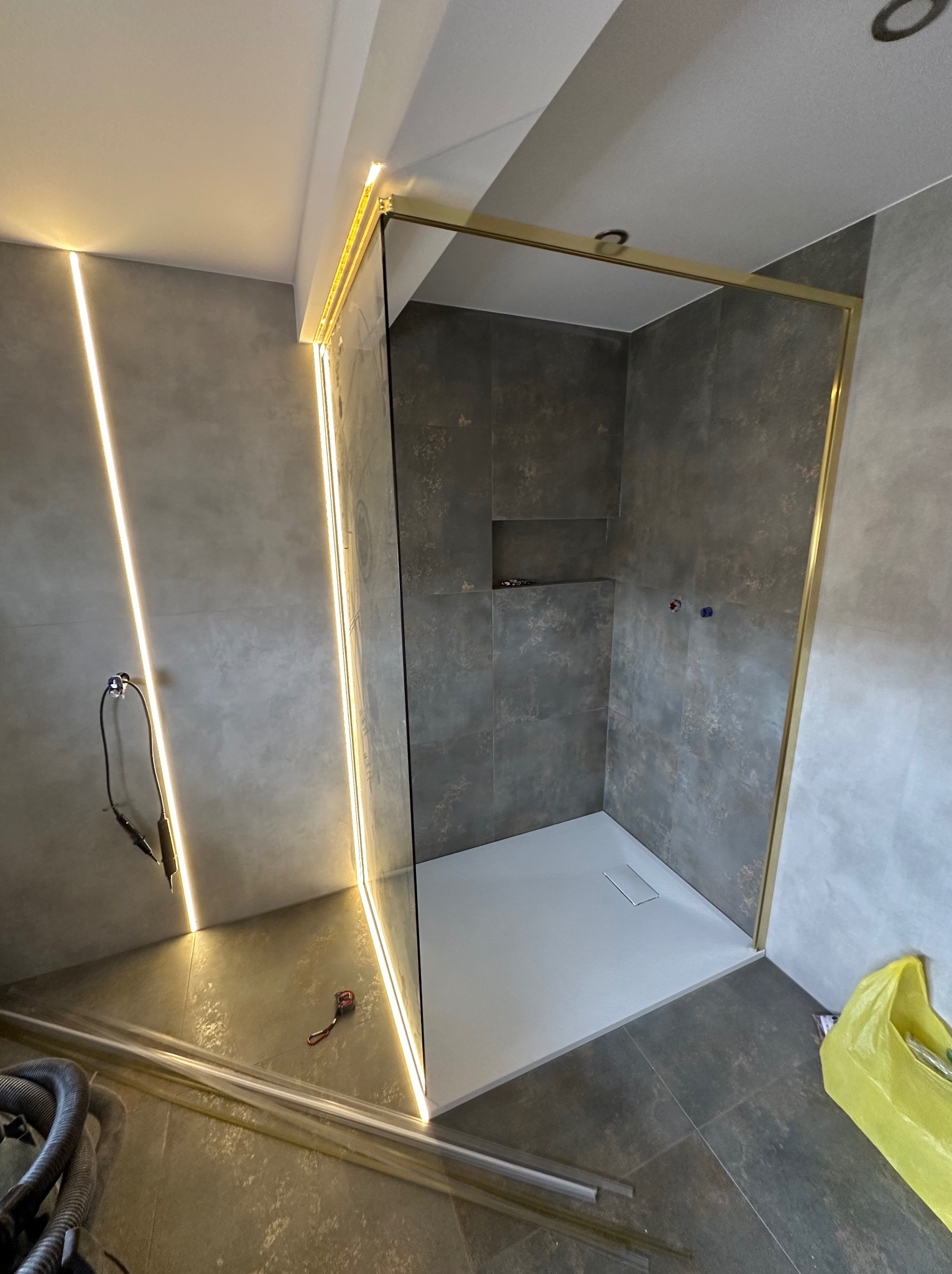 Nowoczesna kabina prysznicowa z brodzikiem i złotymi profilami, podświetlona liniowym oświetleniem LED w szarej łazience. Częściowo widoczne narzędzia.