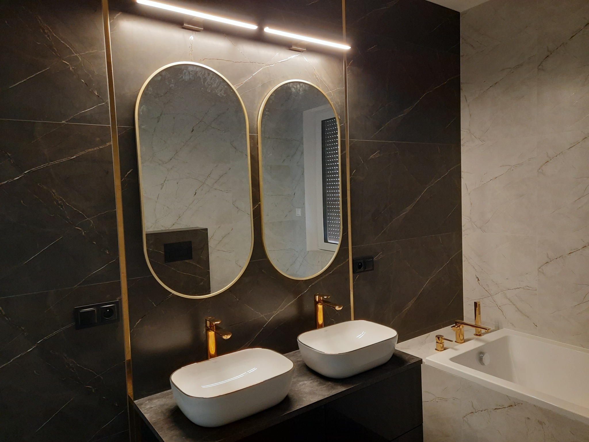Elegancka łazienka z marmurowymi płytkami, dwoma owalnymi umywalkami nablatowymi z złotymi krawędziami i bateriami, dwoma lustrami w złotych ramach i podświetleniem LED