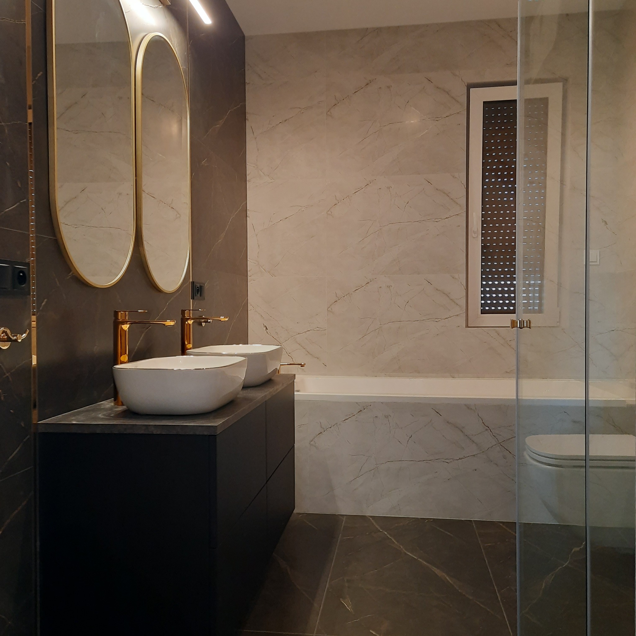 Wykończona łazienka z marmurowymi płytkami, owalnymi lustrami w złotej ramie, dwoma białymi umywalkami nablatowymi z złotymi bateriami, kabiną prysznicową ze szklanymi drzwiami i oknem z roletami.