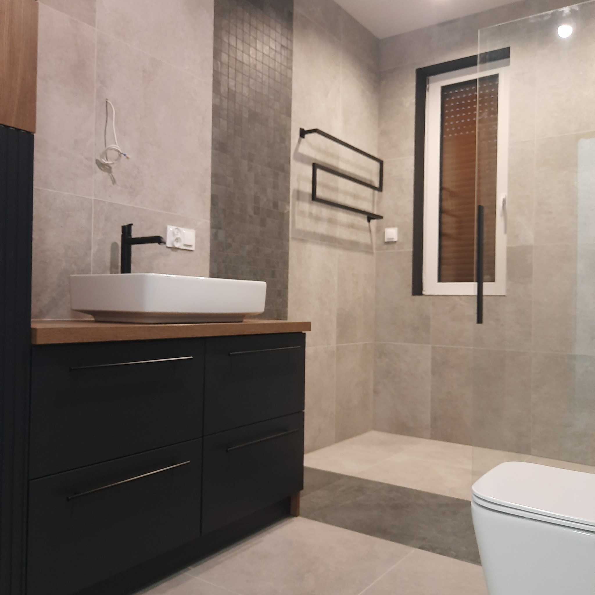 Nowoczesna łazienka z beżowymi płytkami, czarną szafką pod umywalką z drewnianym blatem, wiszącym grzejnikiem drabinkowym i kablami wystającymi ze ściany.